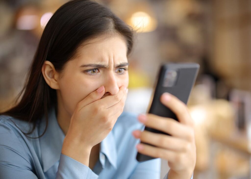 A person in a blue shirt looks at a phone with a surprised expression, covering their mouth. The background is softly blurred.