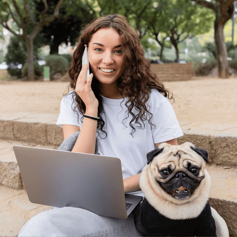 A person with curly hair sits outdoors, using a laptop and phone, accompanied by a pug dog. Trees and brick steps feature in the background.
