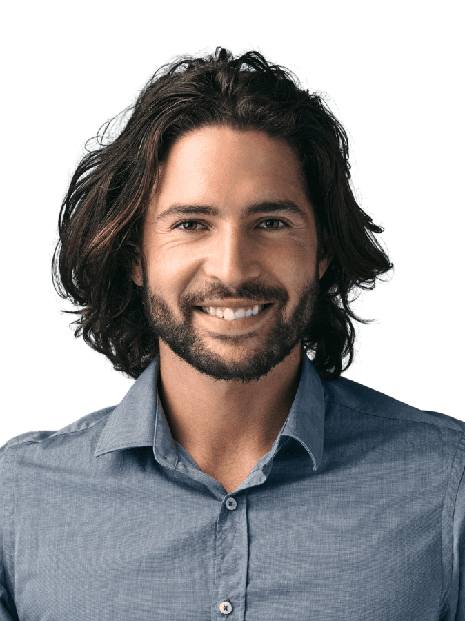 A person with long hair and a beard smiles, wearing a blue shirt. The background is black, creating a portrait-style image.