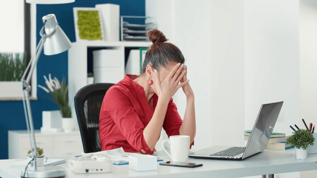 A person sits at a cluttered desk in an office, appearing stressed with a laptop, phone, and a cup nearby.