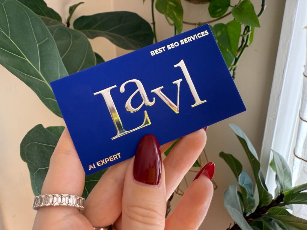 A person with red nails holds a blue card with "Lav1" and "Best SEO Services" near green plants by a window.