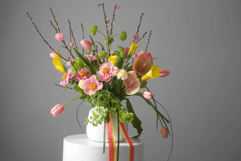 A colorful floral arrangement with pink, yellow, and green flowers is displayed in a white vase on a pedestal against a gray background.