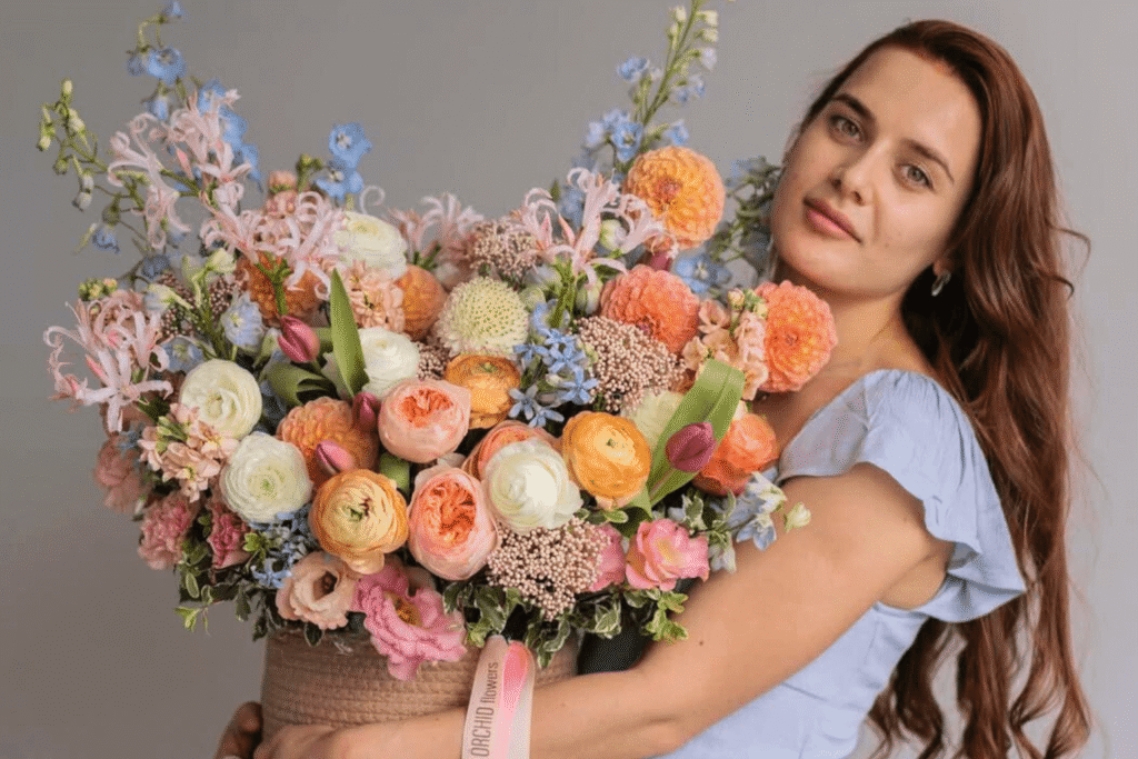 A person with long hair holds a large, colorful bouquet of flowers, featuring pink, orange, and white blooms on a neutral background.