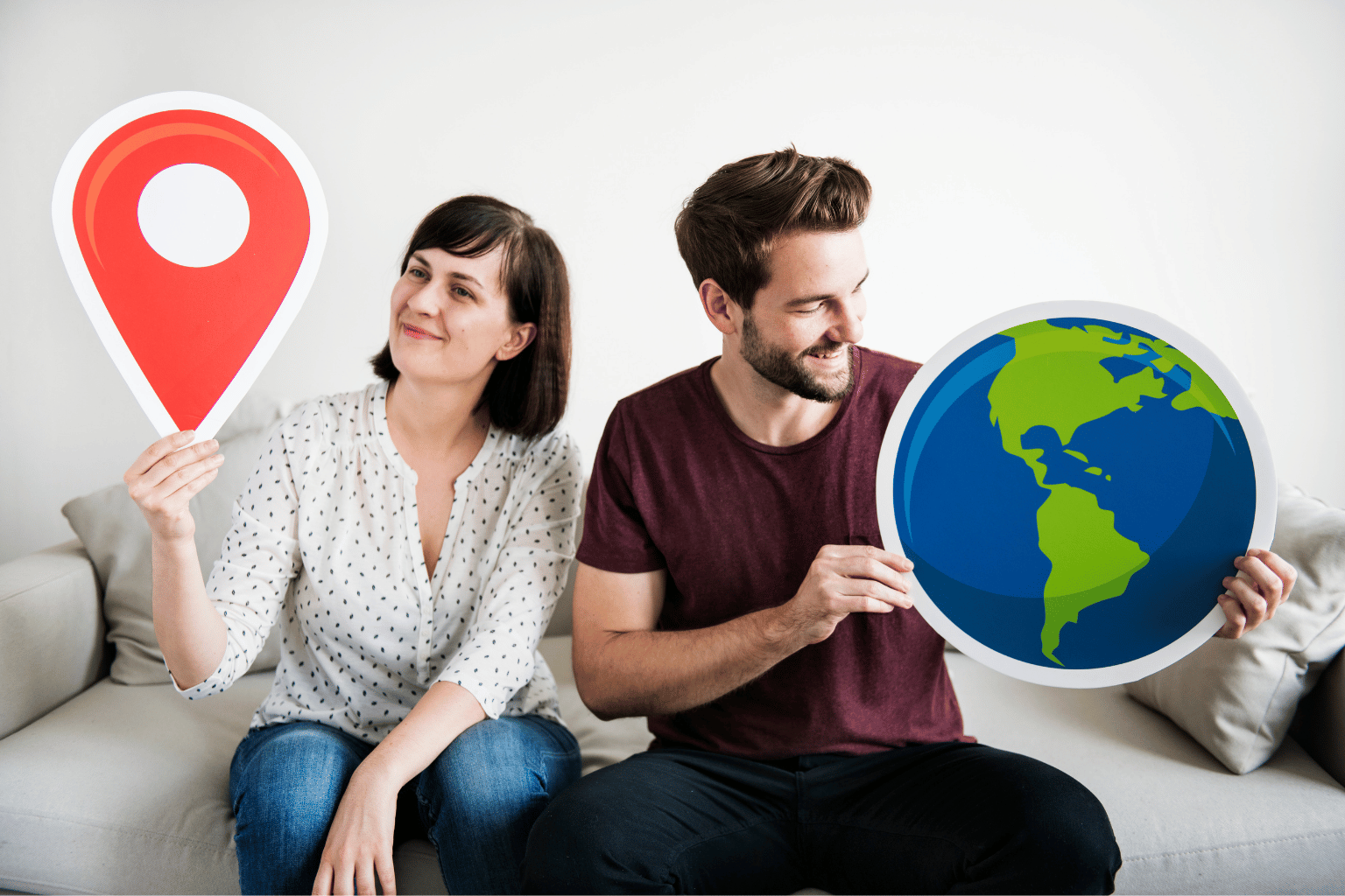 A couple sits on a couch holding a large location pin and a globe.