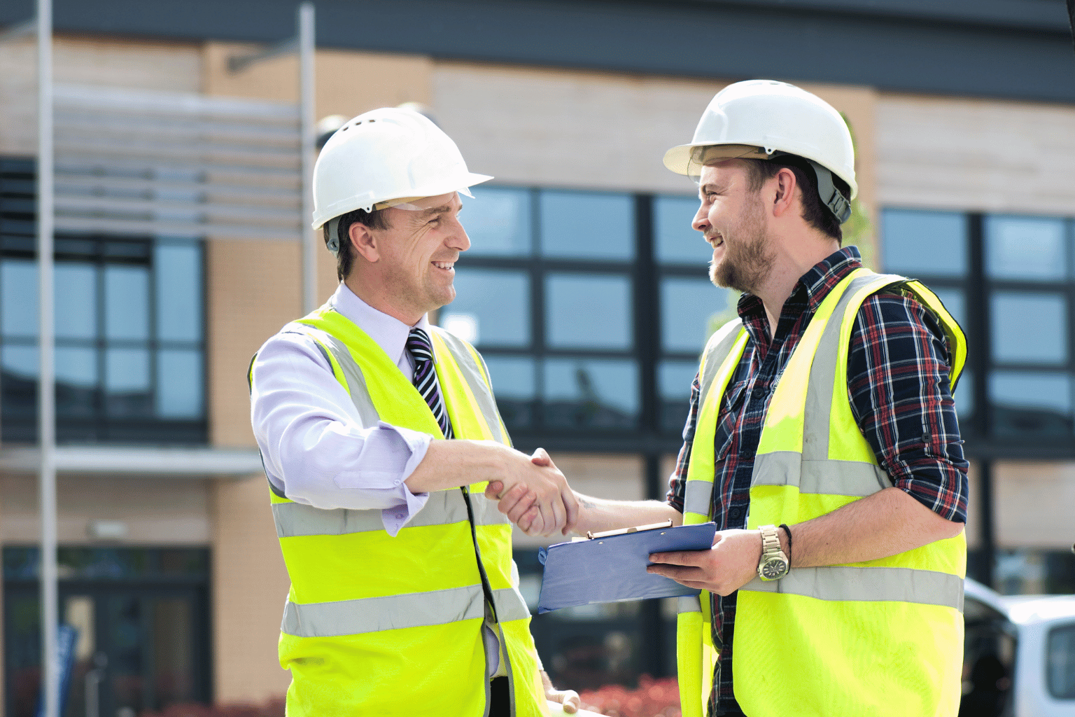 Two construction workers wearing hard hats and high-visibility vests shake hands outside a modern building.