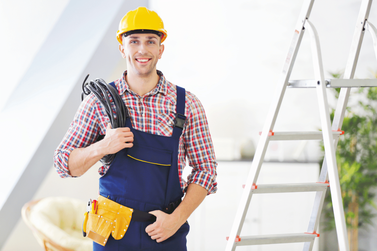 Smiling electrician in a plaid shirt and blue overalls, wearing a yellow hard hat and standing beside a ladder.