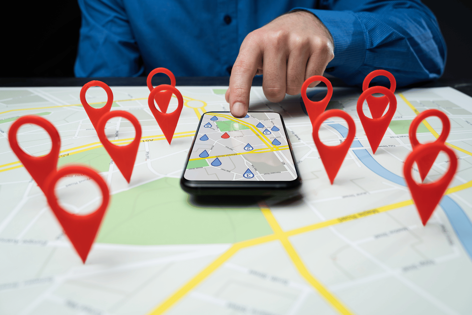 A person in a blue shirt points to a smartphone resting on a printed map marked with red location pins.