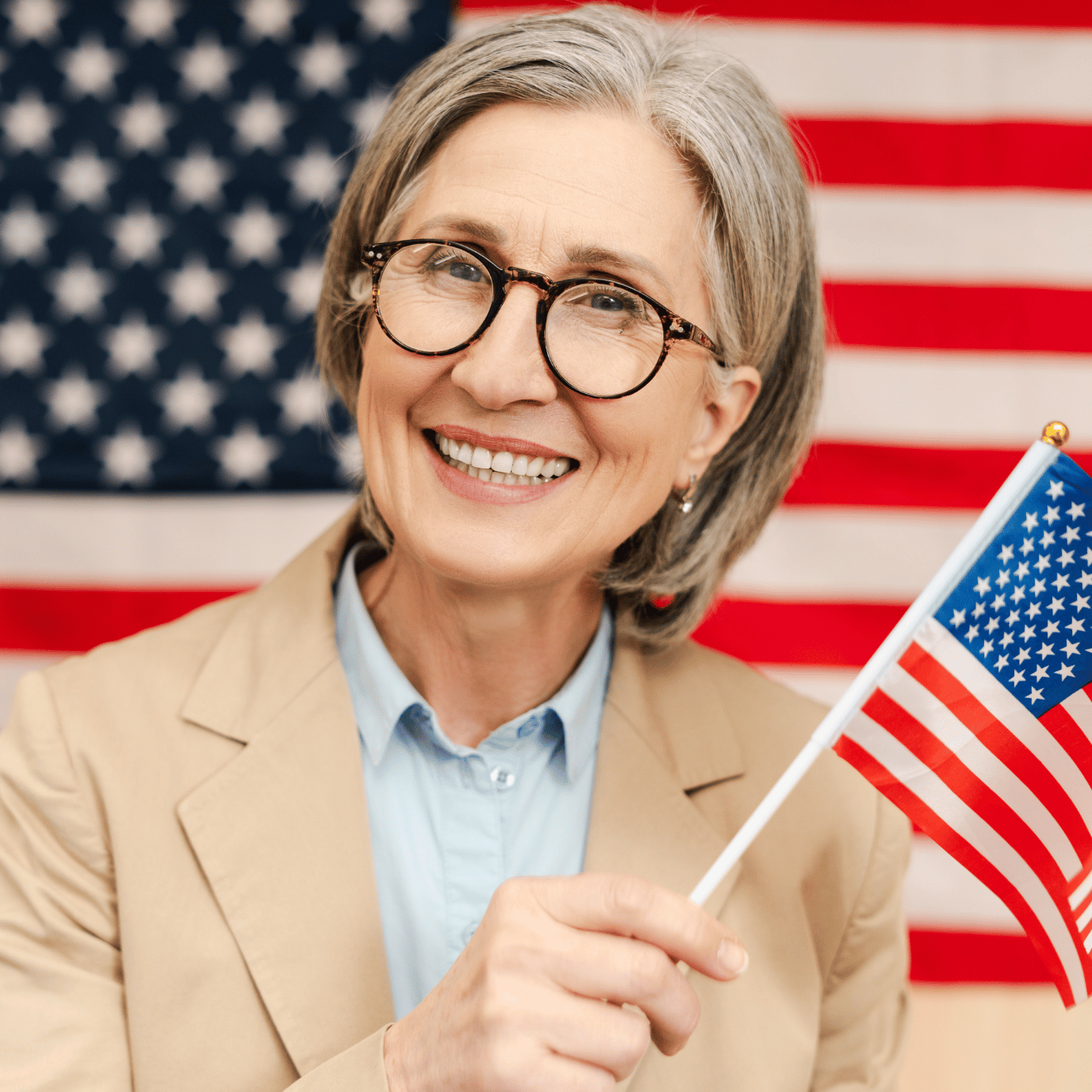 Smiling older woman with glasses in a beige blazer holds a small American flag against a backdrop of a large American flag.